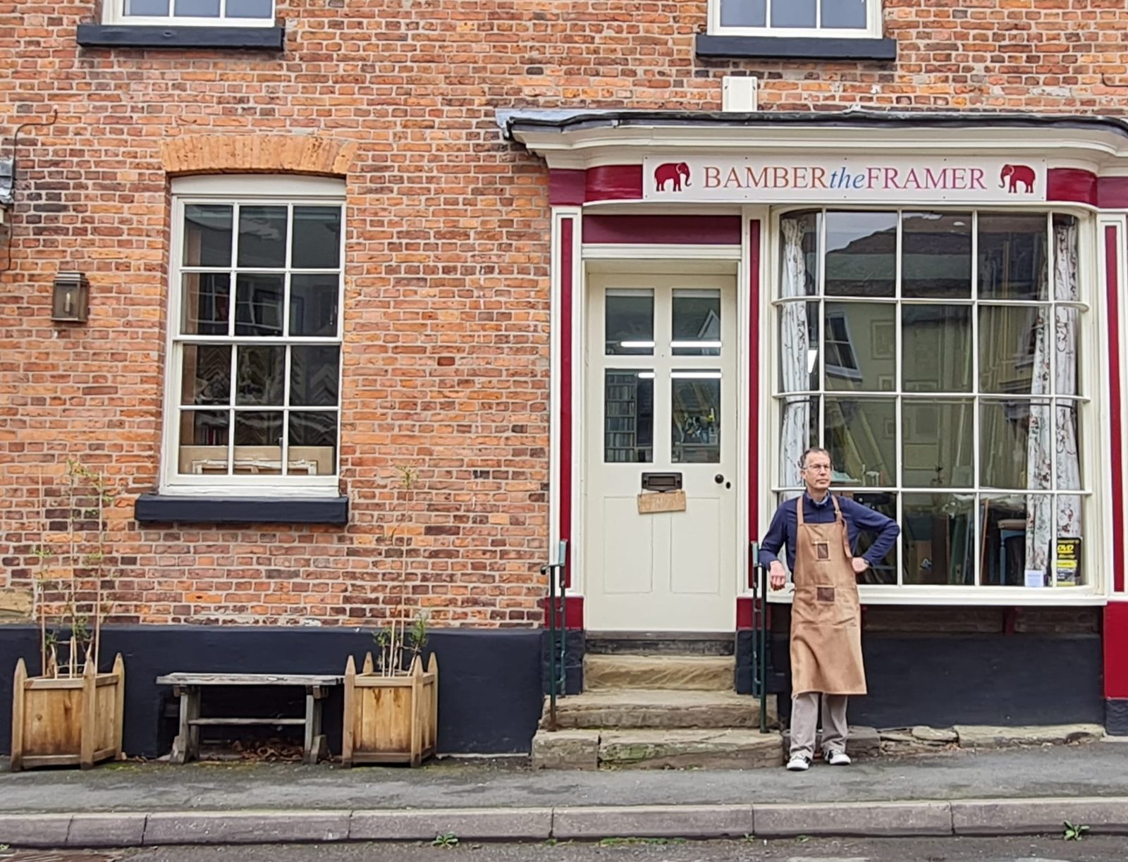 The Bamber the Framer shopfront at 16 Market Square, Bishop's Castle, an oxblood fascia with cream lettering and two red elephants, Bamber standing outside in a leather apron